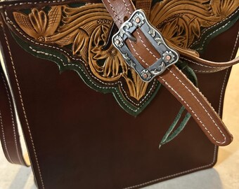 Puede incluir: Un bolso de mano de cuero marrón con intrincados grabados florales y una hebilla plateada decorativa. El bolso presenta una correa de cuero marrón y detalles en verde. Las costuras blancas perfilan los bordes del bolso.