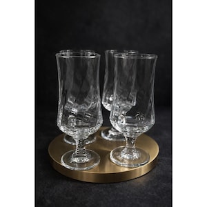 May include: Four clear, stemmed glasses with textured sides are arranged on a round, gold-colored tray. The glasses have a tulip shape and sit on a dark background. The glasses are made of clear glass.