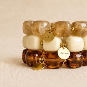 Peut inclure: Un empilement de trois bracelets composés de perles carrées aux bords arrondis dans des tons beige, crème et brun foncé. Les bracelets comportent des breloques en métal doré, dont une gravée J & V et une autre gravée du mot Maman.