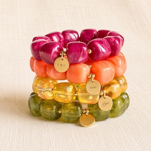 Peut inclure: Une pile de quatre bracelets élastiques composés de perles carrées et épaisses en magenta, orange, jaune et vert olive. Chaque bracelet comporte une petite breloque ronde en métal doré. Les breloques portent les inscriptions L&C, MAMA et Love.