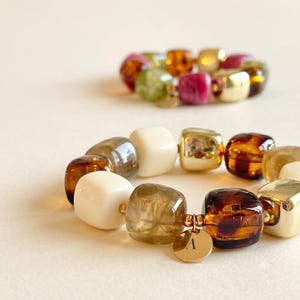 Peut inclure: Deux bracelets extensibles composés de perles carrées dans des tons crème, marron translucide et or métallique. Le bracelet au premier plan comporte une petite breloque ronde en métal doré gravée de la lettre majuscule A.