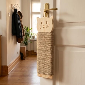 Può includere: Un tiragraffi per porta a forma di gatto in legno con una superficie per graffiare in corda di iuta. La faccia del gatto ha occhi, naso e baffi ritagliati. Il tiragraffi è appeso a una porta con un anello di corda.