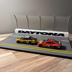 May include: A miniature Daytona racetrack display featuring two stock car models. One yellow car is numbered 20 with DeWalt branding, and one red car is numbered 78 with Bass Pro Shops and Tracker branding. The display includes a gray track surface with yellow lines and a white fence.