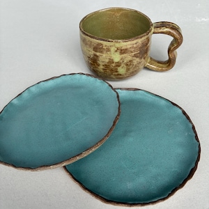May include: A handmade ceramic mug with a unique handle and two matching turquoise plates. The mug has a brown and yellow exterior with a green interior. The plates have a textured surface and a brown rim.