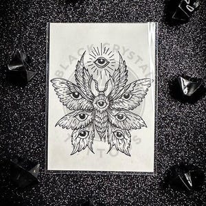 May include: A black and white tattoo design featuring a moth with multiple eyes and wings, with the text "BLACK CRYSTAL TATTOOS". The design is on a white card, surrounded by black crystals on a glittery black surface.