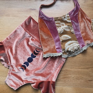 Puede incluir: Un conjunto de terciopelo de dos piezas en tonos melocotón, naranja y morado. La parte superior tiene un estampado floral y ribete de encaje. Los pantalones cortos presentan un diseño de fases lunares.