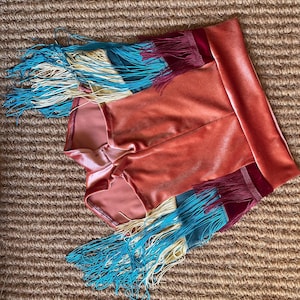 May include: Coral velvet shorts with a high waistband and colourful fringe detailing. The fringe includes shades of turquoise, cream, and burgundy. The shorts are displayed on a textured, neutral-toned surface.