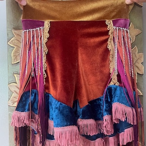 Puede incluir: Una falda de terciopelo de talle alto con una ancha cinturilla dorada, una sección superior de color óxido y un nivel inferior azul oscuro. La prenda está adornada con filas verticales de flecos de pedrería plateada y borlas rosas y burdeos.