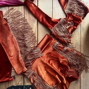 May include: A rust-coloured velvet two-piece set consisting of a halter top and a skirt, both adorned with long, matching fringe trim. The halter top features tie-neck straps with a deep magenta underside, while the skirt has a layered fringe design.