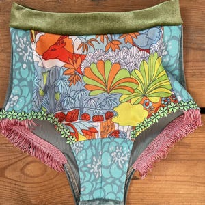 May include: High-waisted underwear with a vibrant floral and animal print. The design features a deer, orange, green, and yellow foliage, and a light blue background. The waistband is green velvet, and the edges are trimmed with pink fringe and green floral trim.