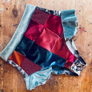 Op de afbeelding: Een patchwork kledingstuk met vierkanten van rode, groene, oranje en bordeaux fluweel en glitterstof. De randen zijn afgewerkt met lichtblauwgroen fluweel. Het kledingstuk heeft een luipaardprint aan de binnenkant.