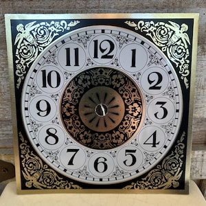 May include: A square clock face featuring a white circular dial with black Arabic numerals from one to twelve. The center has a dark circular pattern with gold accents. The four corners are decorated with gold and black floral motifs.