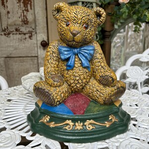 May include: A detailed, antique-style teddy bear statue with a blue bow tie. The bear is a golden brown color and sits atop a green base with gold accents and colorful diamond shapes. The statue is on a white, ornate table.