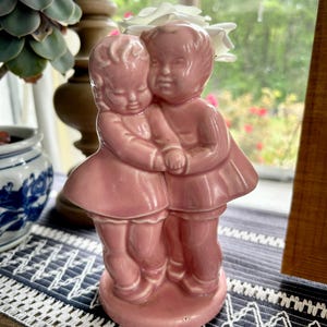 May include: A vintage pink ceramic figurine depicting two embracing children. The figures are dressed in matching outfits and stand on a round base. The figurine has a glossy finish and is placed on a patterned surface.