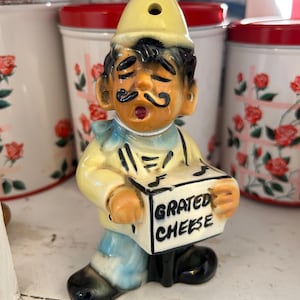 May include: A ceramic figurine of a person wearing a yellow hat and outfit, holding a white box with the text GRATED CHEESE printed in black. The figure has a black mustache, closed eyes, and a blue neck scarf, with black shoes and musical notes.