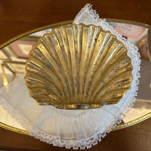 May include: A gold-toned metal dish shaped like a scallop shell with a textured, weathered finish. The shell dish rests on a white fabric doily with a delicate lace trim, which is placed atop an oval-shaped mirror with a thin gold metal frame.