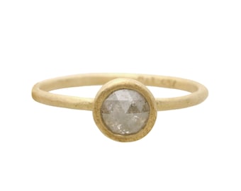 May include: A gold ring with a round, light gray diamond set in a bezel. The band is thin and smooth, and the diamond has a faceted appearance. The ring is set against a white background.