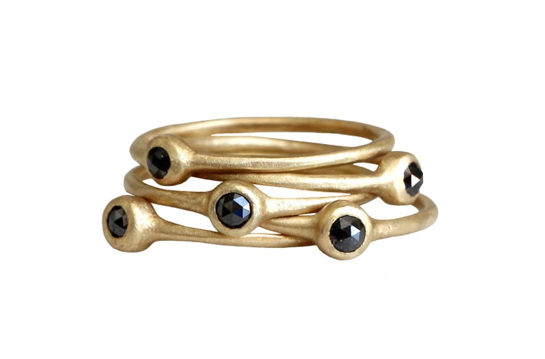 May include: Set of four gold rings with black diamonds. Each ring has a single black diamond set in a smooth gold band.