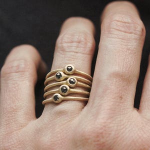 May include: A set of five gold rings with black diamonds stacked on a finger. Each ring has a simple band and a single black diamond.