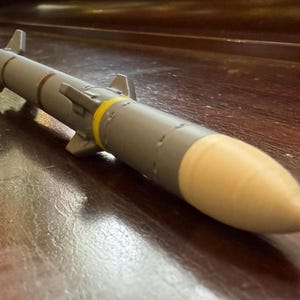 May include: A gray model missile with a tan nose cone and yellow band. The missile has small fins and is resting on a dark wooden surface.