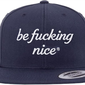 May include: Navy blue snapback hat with the embroidered phrase "be fucking nice" in white script. The hat has a flat brim and a classic six-panel design. The brand logo is visible on the brim.