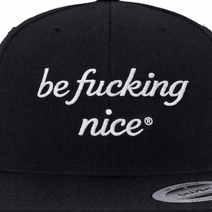 May include: Black snapback hat with the embroidered phrase "be fucking nice" in white cursive lettering. The hat has a flat brim and a structured crown. The hat is a Yupoong brand.