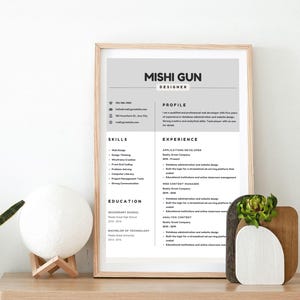 Peut inclure: Un CV encadré avec le nom "Mishi Gun" et le titre "Designer" dans un cadre en bois clair. Le CV comprend des sections pour le profil, les compétences, l'expérience et l'éducation. Une lampe sphérique blanche et une pièce décorative en bois avec de la verdure sont sur la table.