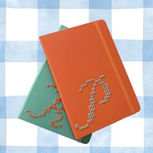 May include: Two notebooks, one orange and one teal, are stacked. The orange notebook features a stitched letter "P", and the teal notebook has a stitched letter "A". Both have an elastic band closure. The background is a blue and white gingham pattern.