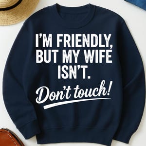 May include: A navy blue sweatshirt featuring the white text: "I'M FRIENDLY, BUT MY WIFE ISN'T. Don't touch!". The sweatshirt has a crew neck and is a solid colour.