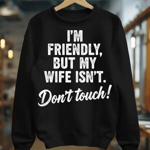 May include: Black sweatshirt with the text "I'M FRIENDLY, BUT MY WIFE ISN'T. Don't touch!" in white. The sweatshirt is hanging on a wooden hanger.