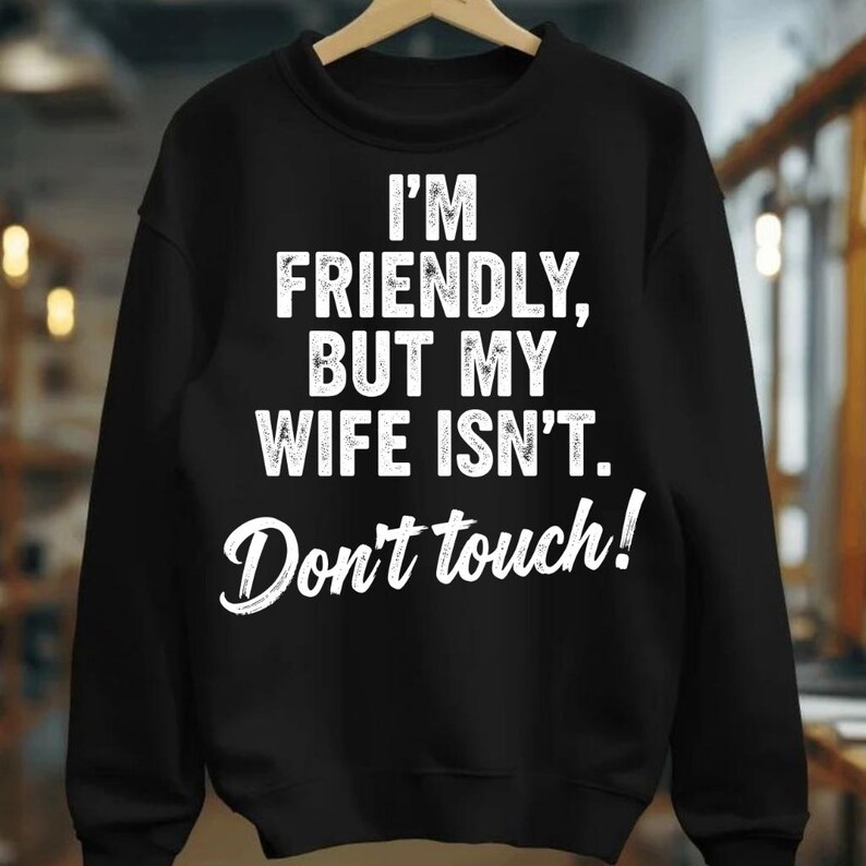 May include: Black sweatshirt with the text "I'M FRIENDLY, BUT MY WIFE ISN'T. Don't touch!" in white. The sweatshirt is hanging on a wooden hanger.