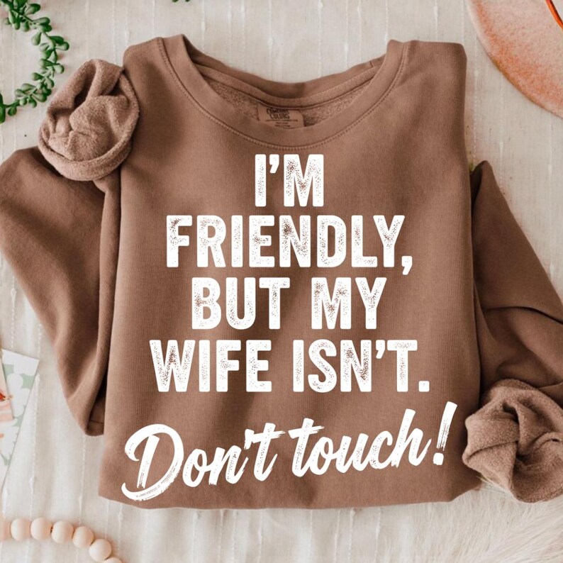 May include: A brown sweatshirt with the text "I'M FRIENDLY, BUT MY WIFE ISN'T. Don't touch!" in white. The sweatshirt is folded, with the sleeves visible. The fabric appears soft and comfortable.