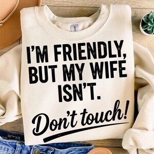 May include: A cream-coloured sweatshirt with the text "I'M FRIENDLY, BUT MY WIFE ISN'T. Don't touch!" in black lettering. The sweatshirt is folded, with blue jeans visible at the bottom. A hat and a small plant are in the background.