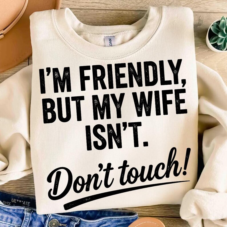 May include: A cream-coloured sweatshirt with the text "I'M FRIENDLY, BUT MY WIFE ISN'T. Don't touch!" in black lettering. The sweatshirt is folded, with blue jeans visible at the bottom. A hat and a small plant are in the background.