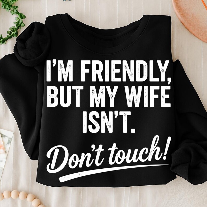 May include: Black sweatshirt with the text "I'M FRIENDLY, BUT MY WIFE ISN'T. Don't touch!" in white. The sweatshirt has long sleeves and a crew neck. The text is in a bold, sans-serif font.