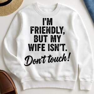 May include: White crew neck sweatshirt with the black text: "I'M FRIENDLY, BUT MY WIFE ISN'T. Don't touch!". The image includes a straw hat and a brown shoe. The sweatshirt is a casual style.