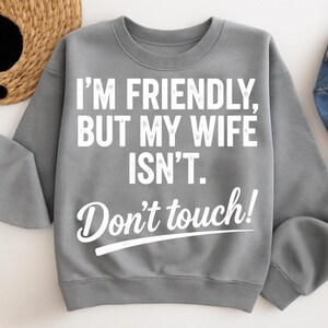 May include: A grey sweatshirt featuring the white text: "I'M FRIENDLY, BUT MY WIFE ISN'T. Don't touch!". The sweatshirt has a crew neck and long sleeves. The text is in a bold, sans-serif font.