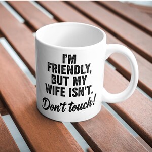 May include: White ceramic mug with a curved handle. The mug features black text that reads: "I'M FRIENDLY, BUT MY WIFE ISN'T. Don't touch!" The mug is sitting on a wooden surface.