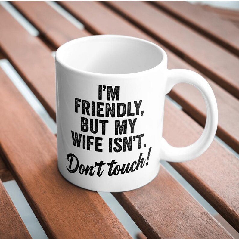 May include: White ceramic mug with a curved handle. The mug features black text that reads: "I'M FRIENDLY, BUT MY WIFE ISN'T. Don't touch!" The mug is sitting on a wooden surface.