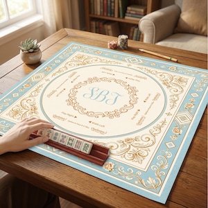 May include: A square mahjong mat with a cream center and light blue border featuring ornate gold floral patterns. The center displays the monogram SBT. The mat includes printed game instructions and directions. A wooden rack holds mahjong tiles.