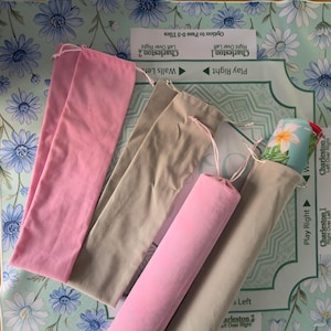 May include: Four long, narrow fabric storage pouches with drawstring closures. Two pouches are solid pink, and two are solid beige. One beige pouch contains a rolled item featuring a light blue background with white and pink floral patterns.
