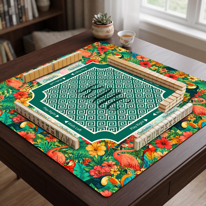May include: A square mahjong mat featuring a vibrant tropical pattern with pink flamingos, colorful parrots, and red hibiscus flowers. The center has a green geometric Greek key border with a monogram, surrounded by mahjong tiles on racks.