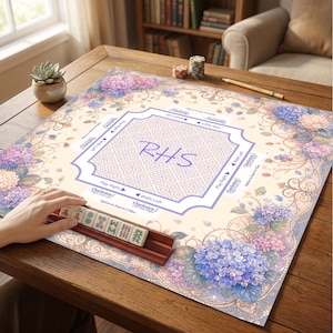 May include: A square Mahjong mat featuring a cream-colored center with a purple Greek key border, the letters RHS, and game instructions. The mat is decorated with blue and purple hydrangea floral patterns and includes a wooden tile rack.