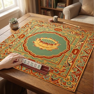 May include: A square mahjong mat featuring a central green lotus flower design surrounded by intricate orange, red, and teal floral patterns. The mat includes the directional labels North, South, East, and West. A wooden tile rack holds several mahjong tiles.