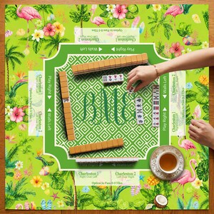 K&ouml;nnte beinhalten: Ein Mahjong-Spielset mit einem gr&uuml;nen Spielbrett, das mit tropischen Flamingos und Hibiskusbl&uuml;ten bedruckt ist. Das Brett enth&auml;lt Anweisungen f&uuml;r Charleston und W&auml;nde, ein zentrales Monogramm und beigefarbene Spielsteine. Eine Teetasse steht bereit.