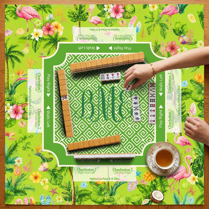 K&ouml;nnte beinhalten: Ein Mahjong-Spielset mit einem gr&uuml;nen Spielbrett, das mit tropischen Flamingos und Hibiskusbl&uuml;ten bedruckt ist. Das Brett enth&auml;lt Anweisungen f&uuml;r Charleston und W&auml;nde, ein zentrales Monogramm und beigefarbene Spielsteine. Eine Teetasse steht bereit.