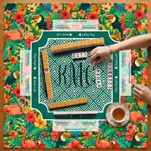 May include: A square mahjong mat featuring a vibrant tropical pattern of pink flamingos, toucans, and red hibiscus flowers. The center has a green geometric design with the letters KMC and several mahjong tiles arranged on top.