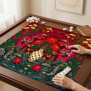 May include: A square mahjong mat featuring a deep red background with two pink tigers, a central cream-colored vase with black dots, red lotus flowers, green foliage, and traditional pagoda structures. White mahjong tiles are scattered on the mat.
