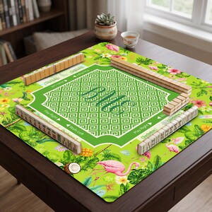 K&ouml;nnte beinhalten: Eine quadratische Mahjong-Matte mit einem leuchtend gr&uuml;nen Tropenmuster aus rosa Flamingos, Hibiskusbl&uuml;ten und Palmbl&auml;ttern. Die Mitte zeigt einen wei&szlig;en geometrischen Rand mit dem Monogramm BME und vier Reihen von Mahjong-Spielsteinen.