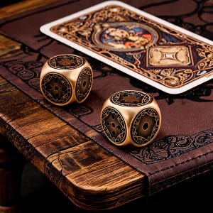 Puede incluir: Dos dados de metal dorado con intrincados patrones de filigrana negra y puntos circulares. Los dados están colocados cerca de una tarjeta rectangular con una ilustración detallada y ornamentada en tonos marrón, oro y azul, sobre una superficie oscura.